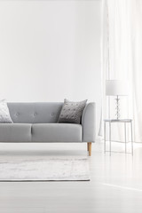 Grey couch next to lamp on silver table in white flat interior with copy space on empty wall. Real photo