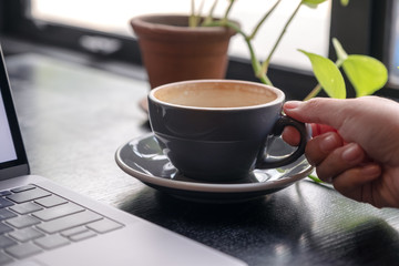 Closeup image of a woman's hand holding a cup of coffee with laptop on wooden table