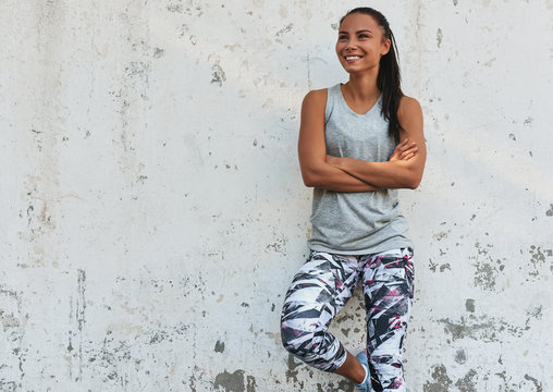 Happy Young Fit Woman Smiling During A Break Outdoors Standing Against Concrete Wall. Athletic Female Resting After Workout Outside.