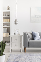 Silver lamp above cabinet next to grey sofa in white living room interior with bookshelf. Real photo