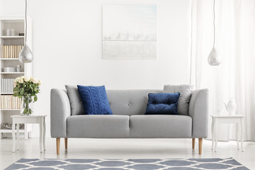 Blue pillows on grey couch next to table with flowers in white flat interior with painting. Real...