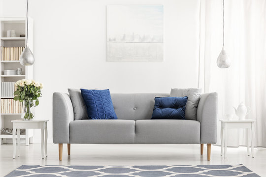 Blue pillows on grey couch next to table with flowers in white flat interior with painting. Real photo