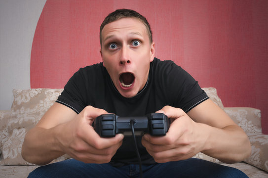 Surprised and enthusiastic gamer man with a joystick in his hands is sitting on a sofa with wide open mouth and is playing in video game. Exciting game concept...