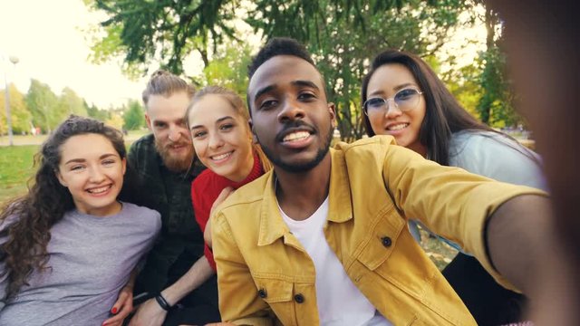 Happy friends are making online video call looking at camera, talking and laughing while African American man is holding device with camera during picnic in park.