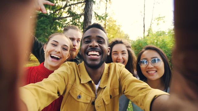Point of view shot of friends taking selfie in park looking at camera, making funny faces and laughing having fun. Modern technology, friendship and nature concept.