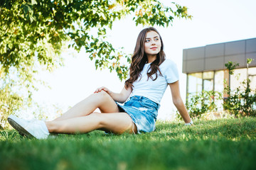 Naklejka na ściany i meble beautiful girl sitting on green grass in park and looking away
