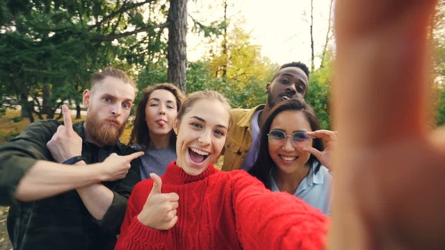 Point of view shot of young woman holding device with camera and taking selfie with friends multi-ethnic group in park in autumn. Photography and people concept.