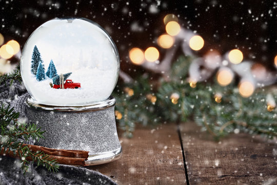 Rustic image of a snow globe with old pick up tuck hauling a Christmas tree surrounded by pine branches, cinnamon sticks and a warm gray scarf with gently falling snow flakes and blurred background.