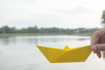 Hand holding yellow paper boat on the lake