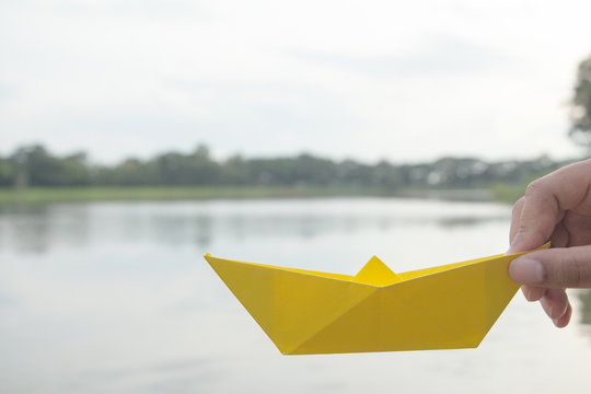 Hand Holding Yellow Paper Boat On The Lake