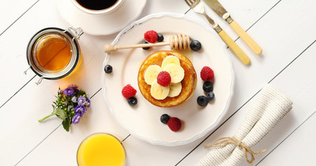 From above shot of nice drinks and cutlery lying on white wooden tabletop near jar of sweet honey and yummy pancakes