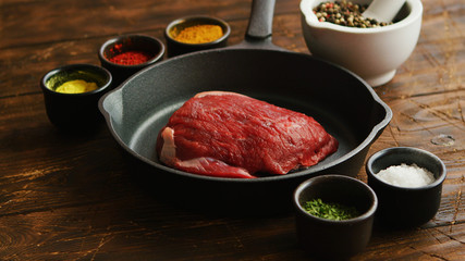 Tiny bowls with aromatic spices standing near frying pan with piece of fresh raw meat on scratched lumber tabletop