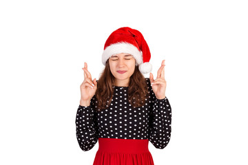 woman withclosing eyes and raising crossed fingers while praying and hoping from dream come true. emotional girl in santa claus christmas hat isolated on white background. holiday concept