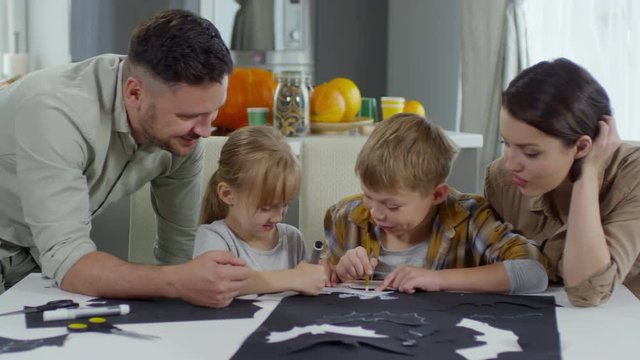 Cute little boy and girl sitting at table in the living room and drawing paper bats with parents while preparing for Halloween