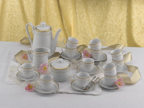Coffee cup set and kettle in white and gold fine porcelain, displayed on a white tablecloth. Yellow curtains in the background