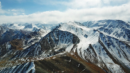 Stone and snow-capped mountains. Blue sky and clouds. Shooting with the drone. Panorama of snowy peaks and rocky mountains. Sharp stones and a gorge are visible.