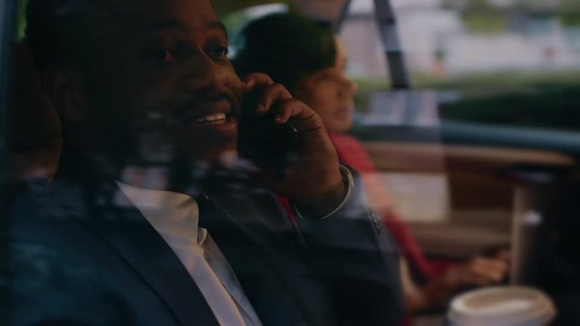 Beautiful Businesswoman and Handsome Businessman Riding on the Backseat of a Car in the Evening. Man Makes a Phone Call, Woman Works on a Laptop. Camera Shot made from Outside the Car.