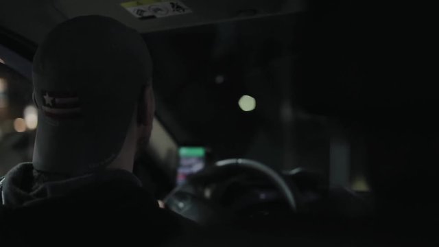 A smiling male driver wearing a cap backwards makes a turn. Driving at night. Back view