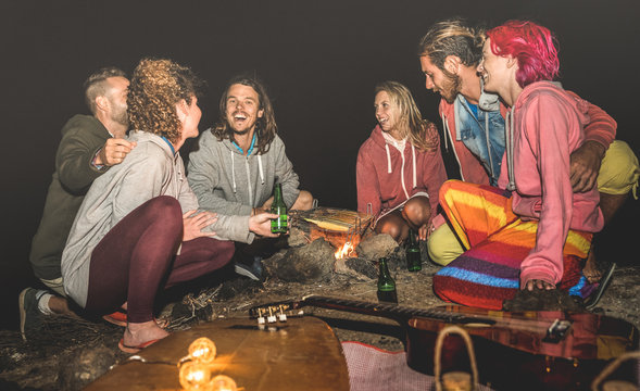 Young hippie friends having fun together at beach camping party with night campfire bbq- Friendship travel concept with young surfer people grilling corn cob at summer barbecue - High iso image