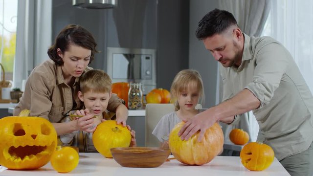 Happy family preparing Halloween decorations in the kitchen: mom and son drawing with pencil on pumpkin and father carving jack-o-lantern with little daughter