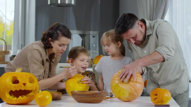 Mother and father carving pumpkins for Halloween with little daughter and son on kitchen table at home