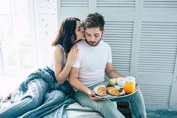 Happy beautiful cute couple in bedroom, they are eating a romantic breakfast in bed while they kissing, hugging and relaxing together. Love, relationships, family and St. Valentines day