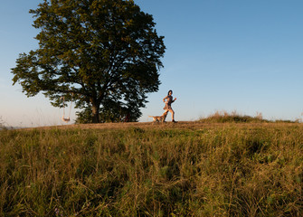 A girl runs with a dog, on a meadow against a big tree background. Best frends.