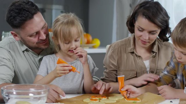 Little girl and boy decorating Halloween cookies with orange pumpkin paste while cooking together with parents on kitchen table