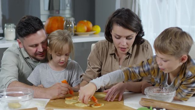 Mother, father, little daughter and son decorating Halloween cookies with orange pumpkin paste while cooking together on kitchen table