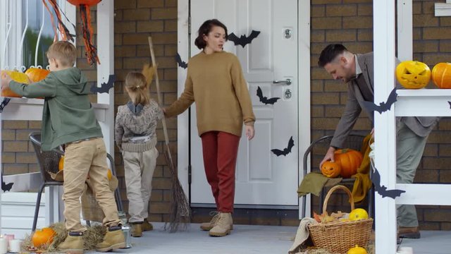 Mother, father and little daughter and son decorating front porch of house with jack-o-lantern and brooms for Halloween
