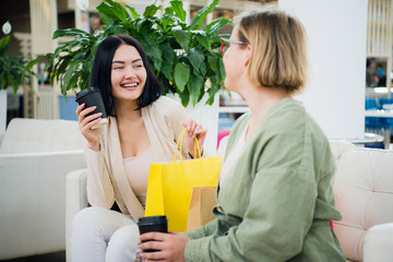 Happy young fashionable women taking a coffee break after shopping, smiling with a coffee-to-go in their hands against shopping mall