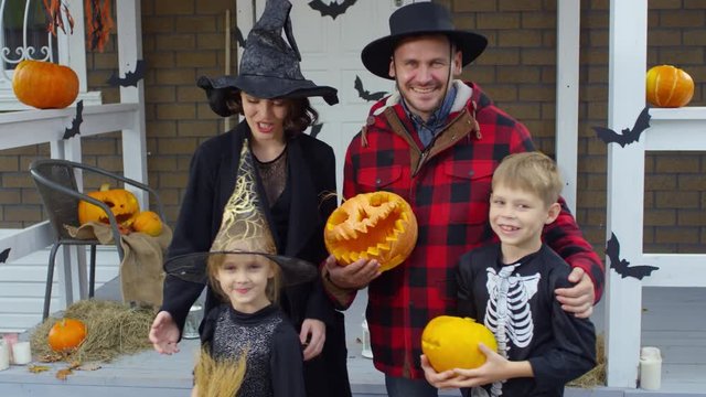 Happy mother, father and two kids in Halloween costumes holding jack-o-lanterns and posing for camera outdoors in front of decorated porch