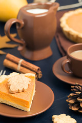 Piece of pumpkin pie with cinnamon and coffee with milk in brown dishes on black background with autumn yellow leaves.
