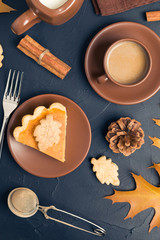 Piece of pumpkin pie with cinnamon and coffee with milk in brown dishes on black background with autumn yellow leaves.