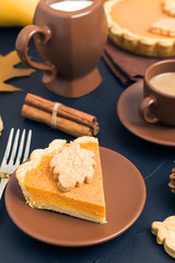 Piece of pumpkin pie with cinnamon and coffee with milk in brown dishes on black background with autumn yellow leaves.