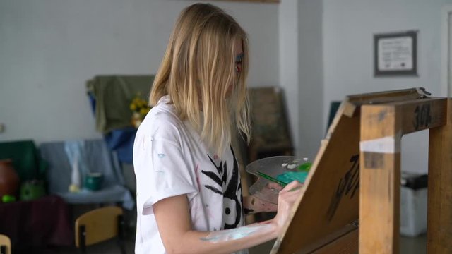 A young artist girl with a face and clothes in the paint, paints a picture on the easel.