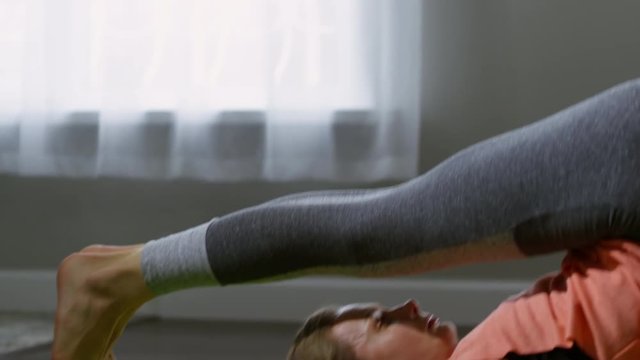 Handheld tracking shot of young woman lying on mat and doing plow pose, then transitioning into shoulderstand while practicing yoga in morning