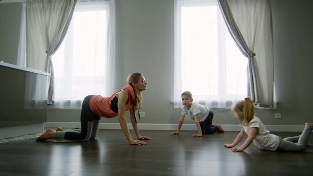 Tracking shot of young mother and children practicing yoga in morning: they are doing cat-cow pose and stretching backs
