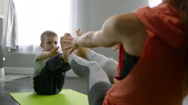 Tracking shot of unrecognizable mother and cute little boy doing yoga pose together: they are touching soles of their feet together and doing forward bend stretch