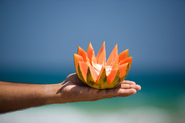  Close up of hand holding beautifully cut in half papaya, with tropical sea backdrop, and blue sky