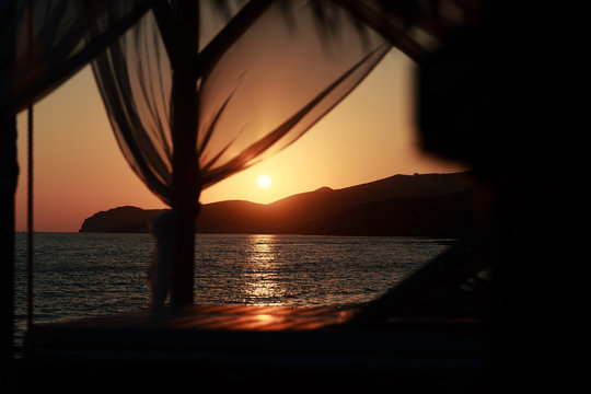 Coucher de soleil vu depuis un lit &agrave; baldaquin avec des reflets sur l'eau et la chaise