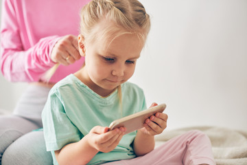 Young mother styling daughter's hair while she's watching cartoons on smart phone