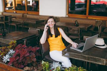 Happy girl in outdoors street cafe sitting at table with laptop pc computer, talking on mobile phone, conducting pleasant conversation, in restaurant during free time. Mobile office freelance concept