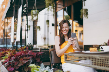 Pretty woman in outdoors street coffee shop cafe sitting at table, listen music in headphones, using mobile phone, relaxing in restaurant on free time. Mobile Office in summer. Lifestyle rest concept.