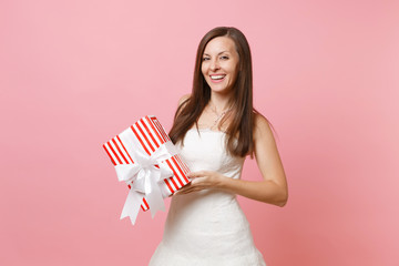 Portrait of smiling tender bride woman in beautiful white wedding dress holding red box with gift present isolated on pink pastel background. Wedding celebration concept. Copy space for advertisement.