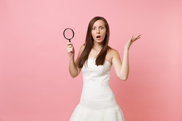Portrait of shocked bride woman in lace white wedding dress spreading hands, holding magnifying glass isolated on pastel pink background. Wedding to do list. Organization of celebration. Copy space.