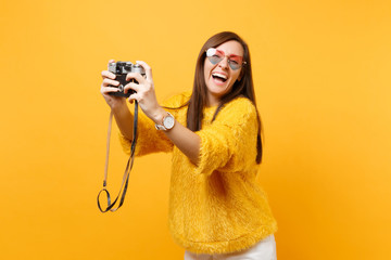 Laughing joyful young woman in heart glasses doing taking selfie shot on retro vintage photo camera isolated on bright yellow background. People sincere emotions, lifestyle concept. Advertising area.