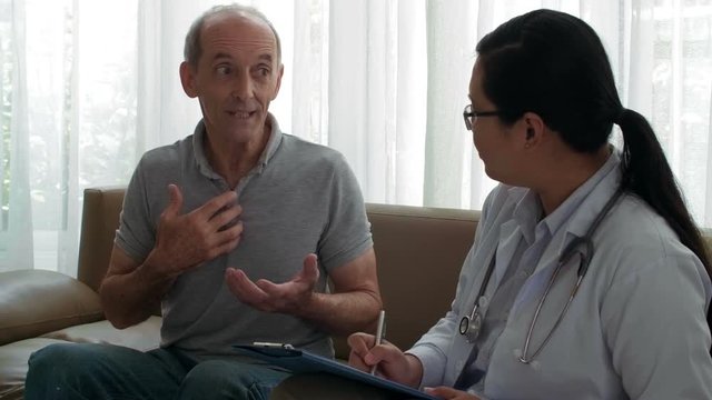 Medium shot of middle-aged Asian man sitting on sofa and talking to attentive female doctor while she is listening to him and writing in document attached to clipboard
