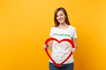 Portrait of woman in white t-shirt with written inscription green title volunteer hold big red wooden heart isolated on yellow background. Voluntary free assistance help, charity grace work concept.