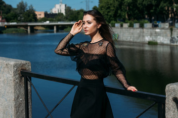 beautiful girl, brunette, in the summer by the river in the city, in a black dress. standing alone by the river, on the bridge, the sun is shining in the face, closed her eyes, rest and tourism.